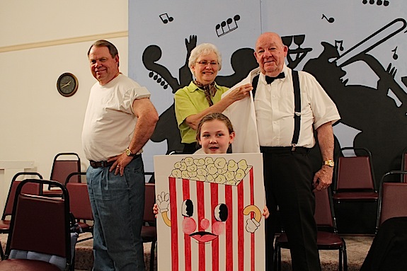 Portraying characters from the movies, the Demopolis Singers will present their spring concert Thursday, May 7. At left Bill Baker is dressed as one of the boys from "Lean on Me." Bobbie Rush, wearing a poodle skirt, helps her husband and director Ed Rush don his white coat as Rick Blaine from "Casa Blanca." Holding the popcorn poster is Meredith Williams.